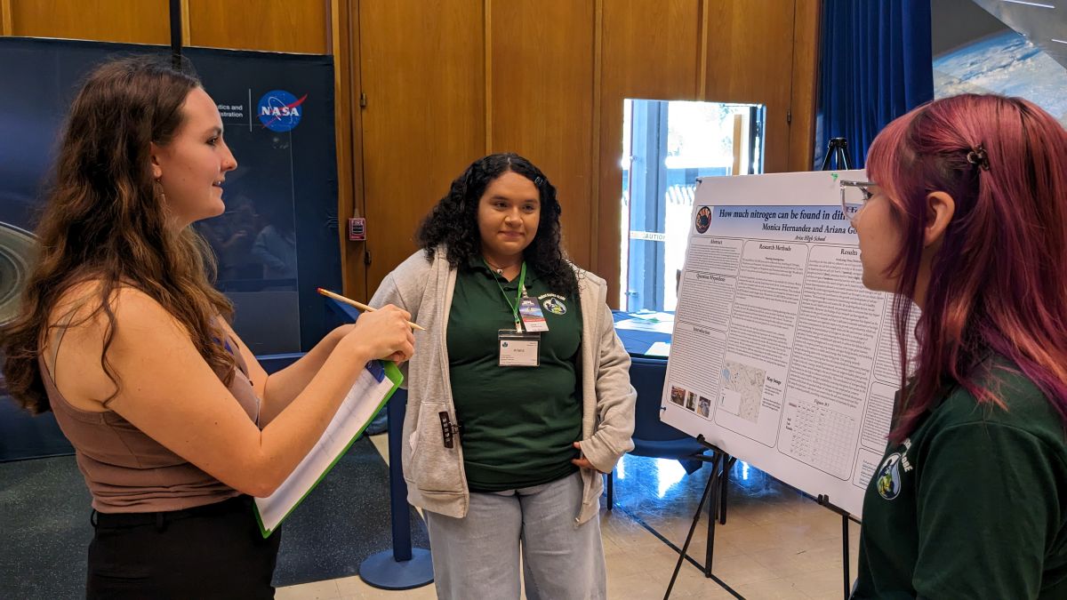 Lexie Gauthier conducts a review of an SRS project, holding a clipboard while two students stand beside their poster